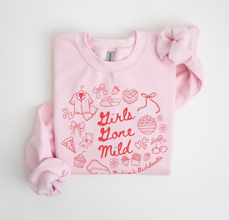 Puede incluir: Sudadera rosa con un dise&ntilde;o gr&aacute;fico rojo que dice "Girls Gone Mild" e incluye varias ilustraciones de art&iacute;culos asociados a una despedida de soltera, como un anillo, una magdalena, una rebanada de pizza y un par de chanclas. El texto en la parte inferior del gr&aacute;fico dice "Madison's Bachelorette."