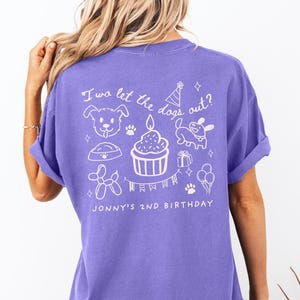 以下が含まれることがあります： 白いイラストとテキストが入った紫色のTシャツ。デザインには犬、カップケーキ、足跡、風船が含まれています。テキストは「I wa let the dogs out？」と「Jonny's 2nd Birthday」と書かれています。