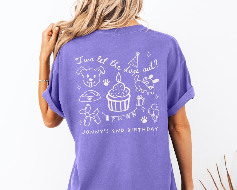 以下が含まれることがあります： 白いイラストとテキストが入った紫色のTシャツ。デザインには犬、カップケーキ、足跡、風船が含まれています。テキストは「I wa let the dogs out？」と「Jonny's 2nd Birthday」と書かれています。