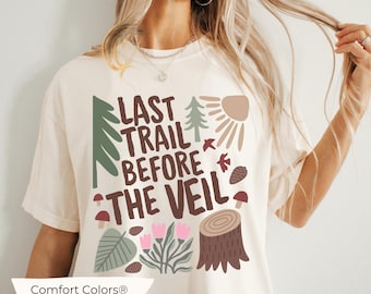 May include: A white t-shirt with a brown graphic design that says "Last Trail Before The Veil". The graphic also includes a tree, a sun, mushrooms, and a tree stump. The shirt is labelled "Comfort Colors® IVORY".