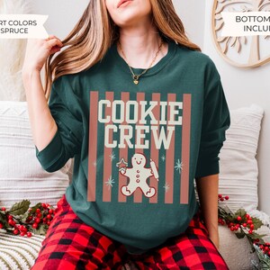 May include: A dark green crewneck sweatshirt featuring a graphic print with vertical muted red stripes and the text COOKIE CREW in cream. Below the text is a gingerbread person holding a rolling pin and a star-shaped cookie cutter.
