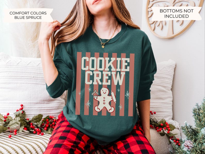 May include: A dark green crewneck sweatshirt featuring a graphic print with vertical muted red stripes and the text COOKIE CREW in cream. Below the text is a gingerbread person holding a rolling pin and a star-shaped cookie cutter.