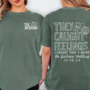 May include: A green t-shirt with white text that reads "THE JACKSONS" and two wedding rings on the front. The back of the shirt says "THEY CAUGHT FEELINGS" with a ring box and disco ball graphic. It also includes "I CAUGHT THIS T-SHIRT the Jackson Wedding 10.05.25".