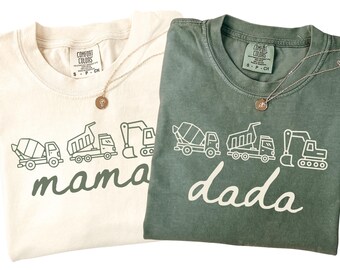 May include: Two crew neck t-shirts, one cream and one sage green, each featuring a graphic of three construction vehicles above the word mama or dada in cursive script. Both shirts have a Comfort Colors label at the inner neckline.
