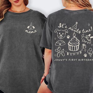 May include: Dark gray t-shirt with a crew neck. The front features a small white illustration of a dog wearing a party hat and the word "mama". The back of the shirt has the text "It's pawty time!" with dog-themed illustrations and "Jonny's First Birthday".