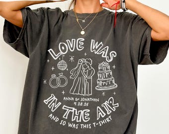May include: A dark gray t-shirt with a white graphic design that says "Love Was In The Air" and includes a wedding cake, a disco ball, and two wedding rings. The text also includes the names "Anna & Jonathan" and the date "7.28.25".