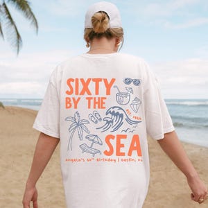 May include: An off-white short-sleeve t-shirt featuring a back graphic with the orange text SIXTY BY THE SEA and blue illustrations of a palm tree, ocean wave, beach chair, umbrella, flip-flops, sunglasses, a drink, and a seashell.