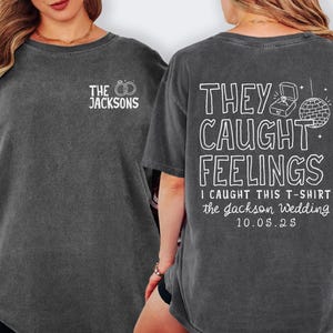 May include: A dark gray t-shirt with white text on the front and back. The front says "THE JACKSONS" with rings. The back says "THEY CAUGHT FEELINGS" with a ring box and disco ball, plus "I CAUGHT THIS T-SHIRT the Jackson Wedding 10.05.25".