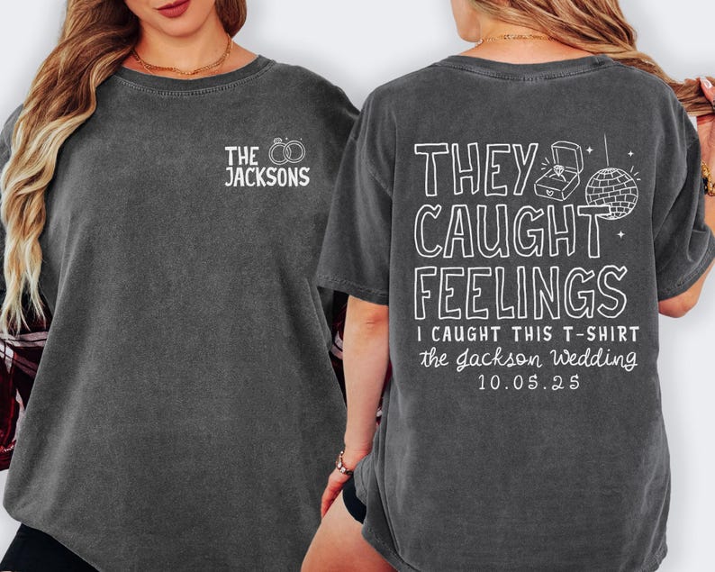 May include: A dark gray t-shirt with white text on the front and back. The front says "THE JACKSONS" with rings. The back says "THEY CAUGHT FEELINGS" with a ring box and disco ball, plus "I CAUGHT THIS T-SHIRT the Jackson Wedding 10.05.25".