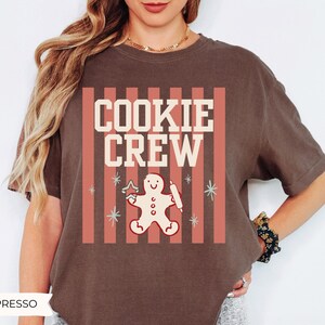 May include: A dark brown short-sleeve t-shirt featuring a graphic print with vertical coral stripes, the text "COOKIE CREW" in cream, and a central illustration of a gingerbread person holding a star-shaped cookie cutter and a rolling pin.
