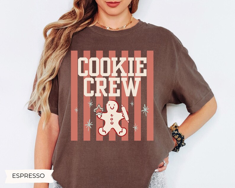 May include: A dark brown short-sleeve t-shirt featuring a graphic print with vertical coral stripes, the text "COOKIE CREW" in cream, and a central illustration of a gingerbread person holding a star-shaped cookie cutter and a rolling pin.