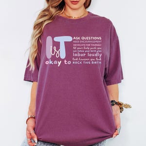 May include: A maroon t-shirt with the words "IT is okay to" and a list of phrases in white, including "ASK QUESTIONS" and "ROCK THIS BIRTH." The text is in a stylized font. The shirt is a loose fit.