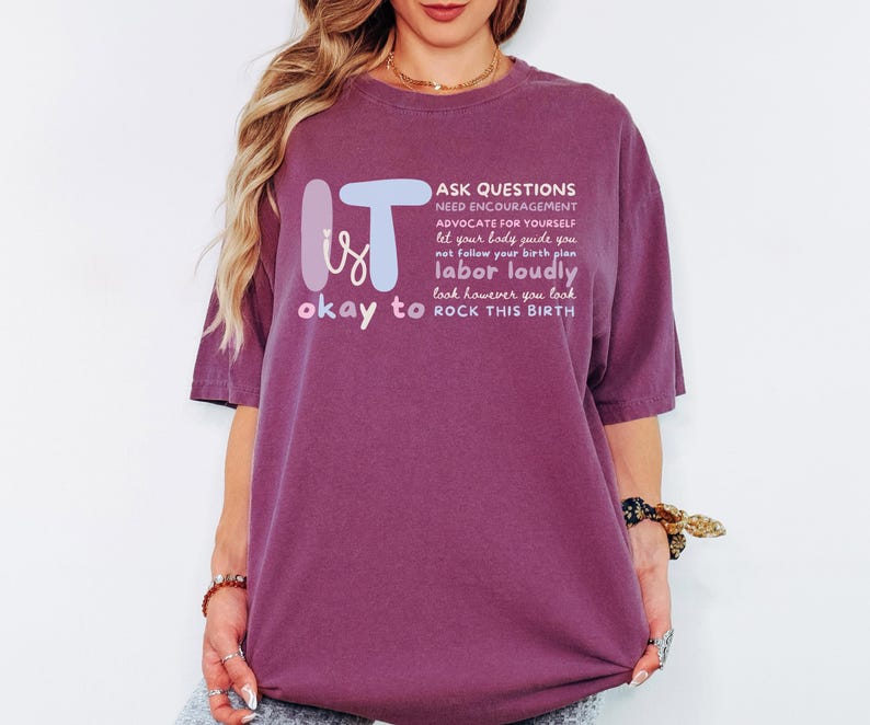 May include: A maroon t-shirt with the words "IT is okay to" and a list of phrases in white, including "ASK QUESTIONS" and "ROCK THIS BIRTH." The text is in a stylized font. The shirt is a loose fit.
