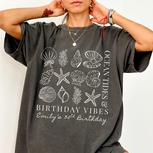May include: Dark gray t-shirt with a white graphic of seashells, starfish, and coral. Text on the shirt reads "OCEAN TIDES & BIRTHDAY VIBES" and "Emily's 30th Birthday". The shirt has short sleeves and a relaxed fit.