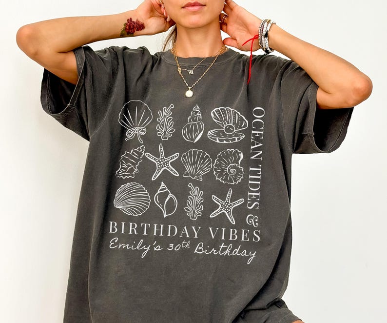 May include: Dark gray t-shirt with a white graphic of seashells, starfish, and coral. Text on the shirt reads "OCEAN TIDES & BIRTHDAY VIBES" and "Emily's 30th Birthday". The shirt has short sleeves and a relaxed fit.