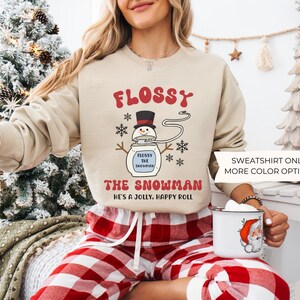 May include: Sand-colored sweatshirt with the text "FLOSSY THE SNOWMAN" and a cartoon snowman design. The sweatshirt also has the text "HE'S A JOLLY, HAPPY ROLL". The model is wearing red and white plaid pajama pants.