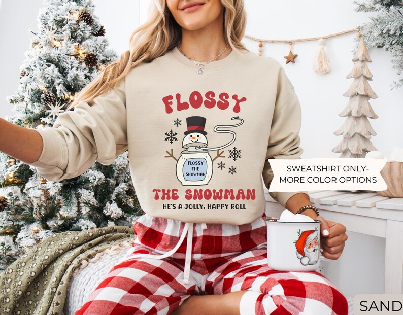May include: Sand-colored sweatshirt with the text "FLOSSY THE SNOWMAN" and a cartoon snowman design. The sweatshirt also has the text "HE'S A JOLLY, HAPPY ROLL". The model is wearing red and white plaid pajama pants.