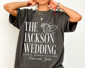 May include: Dark gray t-shirt with white text that reads "THE JACKSON WEDDING" above a ring graphic. Below the text is "ATLANTA, GEORGIA | 8.22.2025" and the names "Anna and Justin". The shirt has short sleeves.