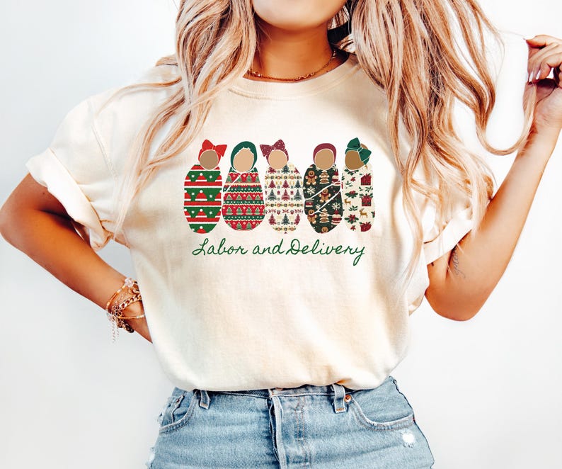 Peut inclure: T-shirt cr&egrave;me avec un motif festif de cinq poup&eacute;es avec des n&oelig;uds, chacune orn&eacute;e de motifs de vacances. Les mots "Labor and Delivery" sont imprim&eacute;s en vert en dessous. Le t-shirt est port&eacute; par une personne.