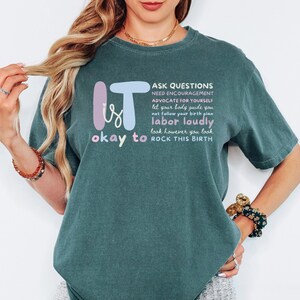 May include: A teal-colored t-shirt with the words "It is okay to" and "Rock This Birth" in a playful font. The shirt also features a list of empowering phrases such as "Ask Questions" and "Labor Loudly."