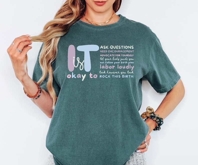 May include: A teal-colored t-shirt with the words "It is okay to" and "Rock This Birth" in a playful font. The shirt also features a list of empowering phrases such as "Ask Questions" and "Labor Loudly."