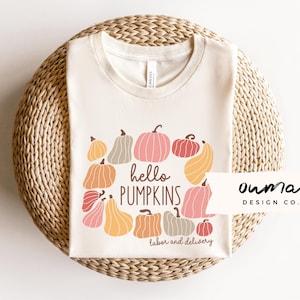 May include: A beige t-shirt with a graphic of pumpkins in various shades of orange, pink, and brown. The text "hello pumpkins labor and delivery" is printed on the shirt. The text "ourma design co." is printed in the bottom right corner.