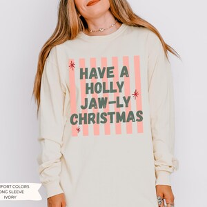 K&ouml;nnte beinhalten: Ein elfenbeinfarbenes Langarmshirt mit einem Grafikdruck aus vertikalen hellrosa Streifen. Der Text lautet HAVE A HOLLY JAW-LY CHRISTMAS in dunkelgr&uuml;nen Blockbuchstaben, akzentuiert durch kleine rote Sternsymbole an den Seiten des Textes.
