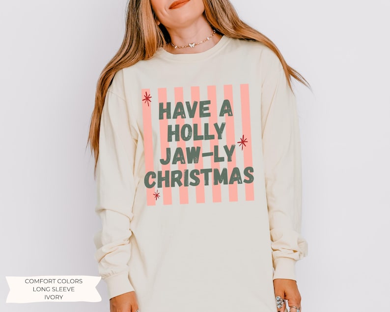 K&ouml;nnte beinhalten: Ein elfenbeinfarbenes Langarmshirt mit einem Grafikdruck aus vertikalen hellrosa Streifen. Der Text lautet HAVE A HOLLY JAW-LY CHRISTMAS in dunkelgr&uuml;nen Blockbuchstaben, akzentuiert durch kleine rote Sternsymbole an den Seiten des Textes.