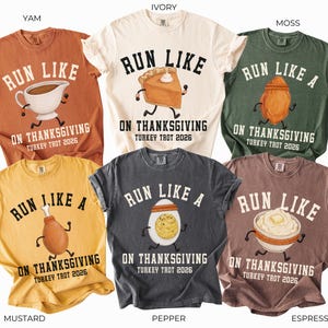 May include: Six short-sleeve crew neck t-shirts in yam, ivory, moss, mustard, pepper, and espresso colors. Each shirt features a graphic of a Thanksgiving food item with arms and legs running, and the text RUN LIKE A ON THANKSGIVING TURKEY TROT 2026.