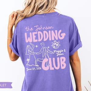 May include: Violet-colored t-shirt with the text "the Johnson WEDDING CLUB" in pink. The design includes a dancing couple, a disco ball, and the date "June 27, 2026." The shirt is a casual style.