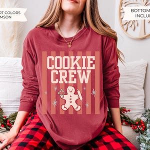 May include: A crimson long-sleeve crewneck sweatshirt featuring a graphic print with the text COOKIE CREW in cream letters. The design includes a gingerbread person holding a rolling pin and a star-shaped cookie, set against vertical stripes.