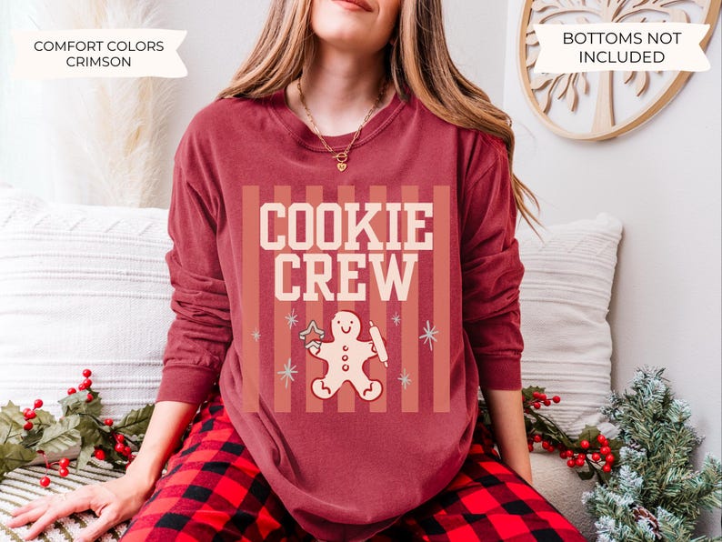 May include: A crimson long-sleeve crewneck sweatshirt featuring a graphic print with the text COOKIE CREW in cream letters. The design includes a gingerbread person holding a rolling pin and a star-shaped cookie, set against vertical stripes.