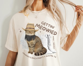 May include: A cream-colored t-shirt with the text "Gettin' Meowied" in brown lettering. The shirt features a cartoon cat wearing a cowboy hat and bandana, and a pair of white cowboy boots. The text "Hannah's Bach Party + Austin, TX" and "Est. 2023" are also printed on the shirt.