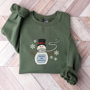 May include: A green sweatshirt featuring a white snowman graphic. The snowman is wearing a black hat and has a blue mouth. The text "Flossy the Snowman" is printed below the snowman.