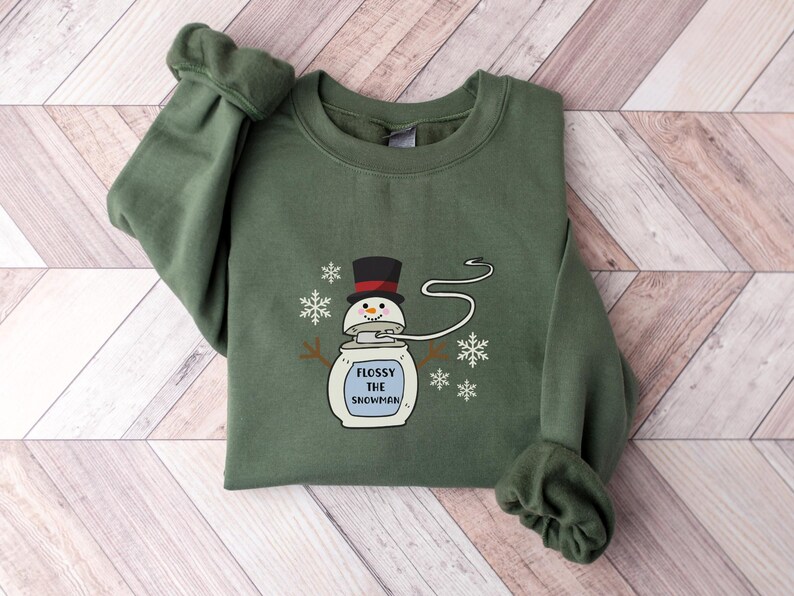 May include: A green sweatshirt featuring a white snowman graphic. The snowman is wearing a black hat and has a blue mouth. The text "Flossy the Snowman" is printed below the snowman.