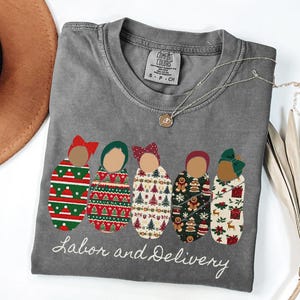 Peut inclure: T-shirt gris &agrave; col rond avec un motif de cinq b&eacute;b&eacute;s emmaillot&eacute;s avec des motifs et des n&oelig;uds diff&eacute;rents. Le texte "Labor and Delivery" est brod&eacute; en dessous. Un collier dor&eacute; et un chapeau marron sont &eacute;galement visibles.