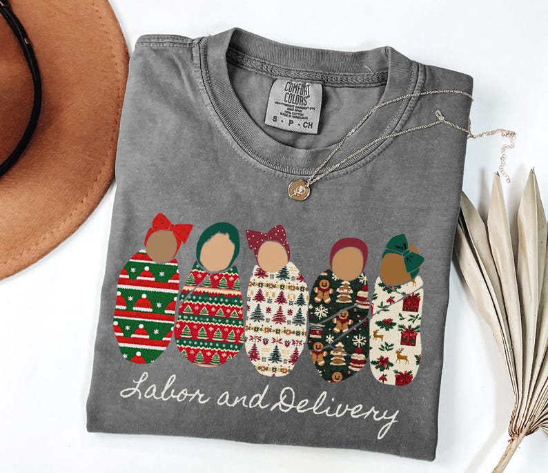 Peut inclure: T-shirt gris &agrave; col rond avec un motif de cinq b&eacute;b&eacute;s emmaillot&eacute;s avec des motifs et des n&oelig;uds diff&eacute;rents. Le texte "Labor and Delivery" est brod&eacute; en dessous. Un collier dor&eacute; et un chapeau marron sont &eacute;galement visibles.