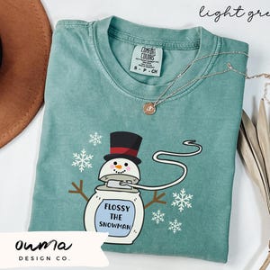 May include: A light green t-shirt with a cartoon snowman holding a roll of floss. The snowman is wearing a black top hat with a red band and has a white face with black eyes and a black mouth. The text "Flossy the Snowman" is printed on the snowman's body. The shirt has a distressed look and is made of a soft, comfortable material.
