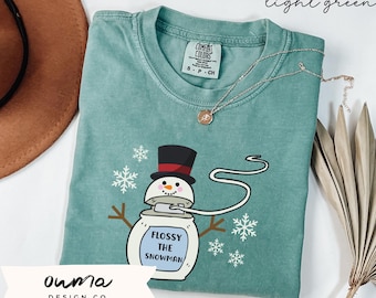 Könnte beinhalten: Ein hellgrünes T-Shirt mit einem Cartoon-Schneemann, der eine Rolle Zahnseide hält. Der Schneemann trägt einen schwarzen Zylinder mit rotem Band und hat ein weißes Gesicht mit schwarzen Augen und einem schwarzen Mund. Der Text "Flossy the Snowman" ist auf den Körper des Schneemanns gedruckt. Das Shirt hat einen Used-Look und ist aus einem weichen, bequemen Material gefertigt.