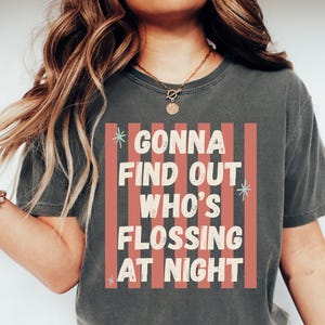 K&ouml;nnte beinhalten: Ein anthrazitfarbenes Kurzarm-T-Shirt mit einem Grafikdruck aus vertikalen korallenfarbenen Streifen und dem Text "GONNA FIND OUT WHO'S FLOSSING AT NIGHT" in cremefarbenen Buchstaben, akzentuiert durch kleine hellblaue sternf&ouml;rmige Symbole.