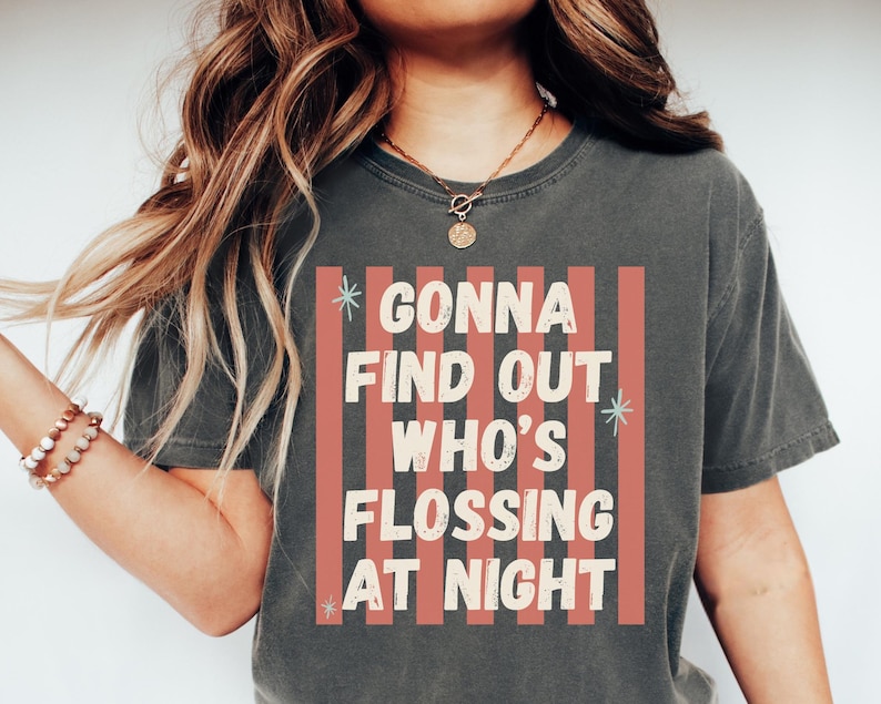 K&ouml;nnte beinhalten: Ein anthrazitfarbenes Kurzarm-T-Shirt mit einem Grafikdruck aus vertikalen korallenfarbenen Streifen und dem Text "GONNA FIND OUT WHO'S FLOSSING AT NIGHT" in cremefarbenen Buchstaben, akzentuiert durch kleine hellblaue sternf&ouml;rmige Symbole.