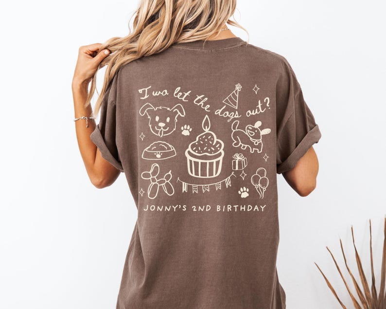 以下が含まれることがあります： 白のグラフィックデザインが施された茶色のTシャツ。デザインには犬、足跡、カップケーキ、風船、そして「I wa let the dogs out?」と「Jonny's 2nd Birthday」の文字が含まれています。このシャツは半袖で、ゆったりとしたフィット感です。