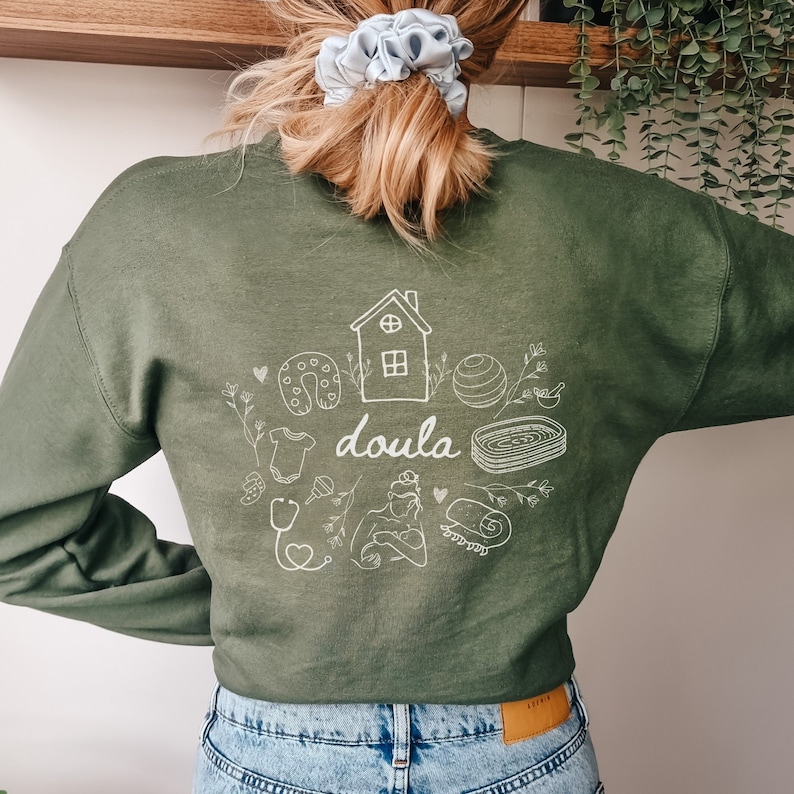 May include: A green sweatshirt with a white graphic design of a house, a baby, a stethoscope, a yoga ball, a horseshoe, and the word "doula".