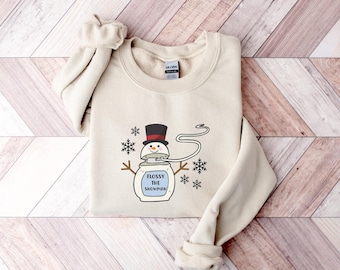 May include: A beige sweatshirt with a cartoon snowman wearing a black top hat and holding a sign that says "Flossy the Snowman".