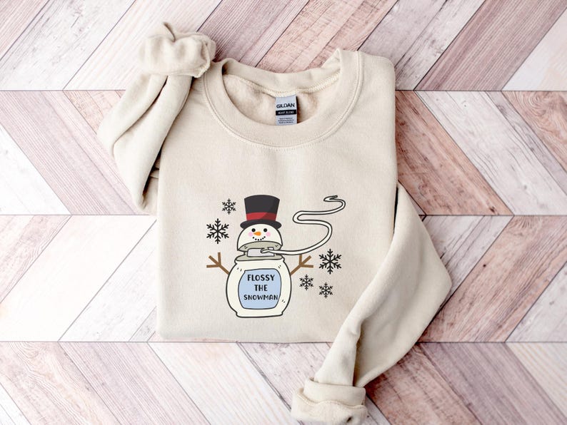 May include: A beige sweatshirt featuring a cartoon snowman wearing a black top hat and holding a sign that reads "Flossy the Snowman".