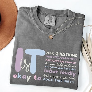 May include: A gray t-shirt with the text "It is okay to" and "Ask questions, need encouragement, advocate for yourself, labor loudly, rock this birth." A gold necklace with a pendant is on the shirt.