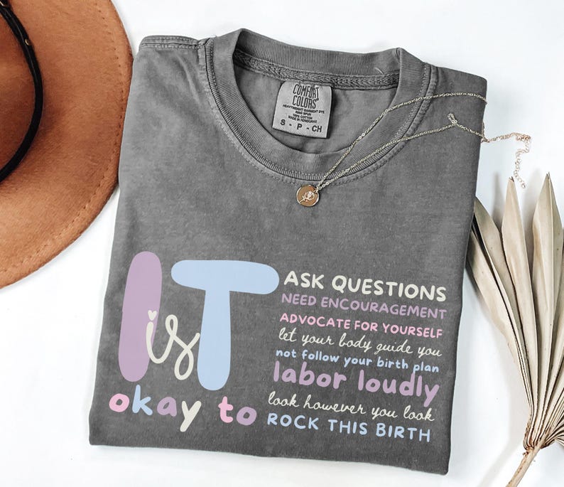 May include: A gray t-shirt with the text "It is okay to" and "Ask questions, need encouragement, advocate for yourself, labor loudly, rock this birth." A gold necklace with a pendant is on the shirt.