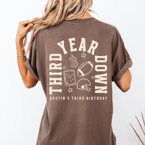 May include: Brown t-shirt with a graphic design on the back. The design includes the words "THIRD YEAR DOWN" and "AUSTIN'S THIRD BIRTHDAY" with football-themed illustrations. The shirt has short sleeves and a relaxed fit.