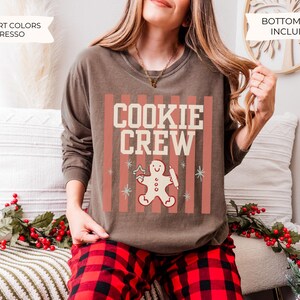 May include: An espresso-colored long-sleeve crewneck sweatshirt featuring a graphic print with vertical salmon-colored stripes, the text "COOKIE CREW" in cream, and a central illustration of a gingerbread person holding a star and rolling pin.