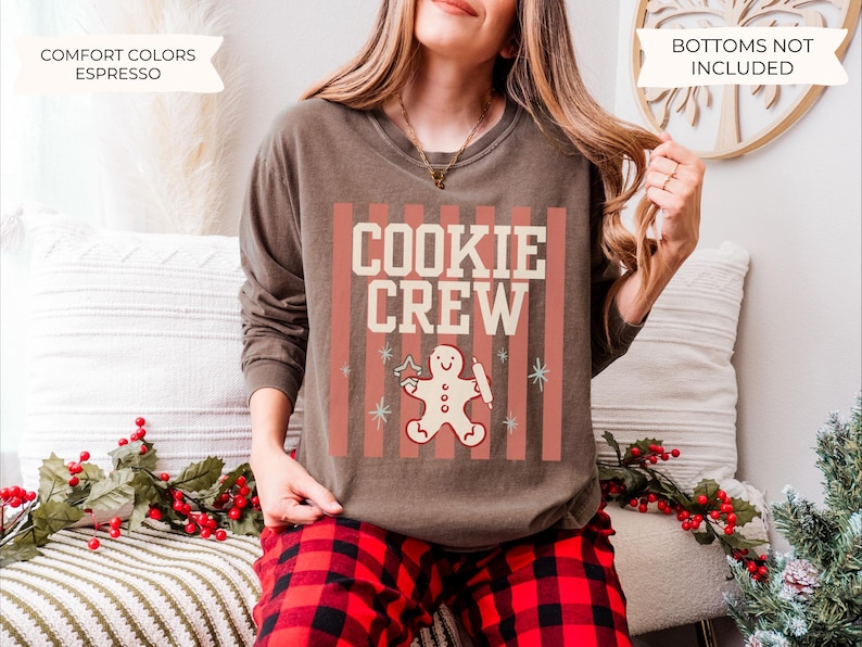 May include: An espresso-colored long-sleeve crewneck sweatshirt featuring a graphic print with vertical salmon-colored stripes, the text "COOKIE CREW" in cream, and a central illustration of a gingerbread person holding a star and rolling pin.